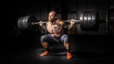 Muscular man with tattoos squats with heavy barbell in gym.