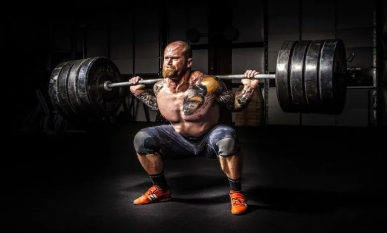 Muscular man with tattoos squats with heavy barbell in gym.