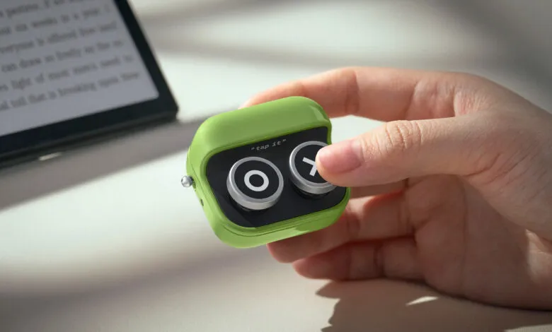 Hand holding green 'tap it' device with circle and cross buttons.