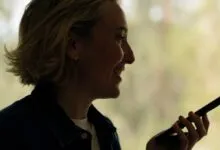Smiling woman with short blonde hair holds a smartphone, silhouetted against a bright window.