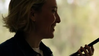 Smiling woman with short blonde hair holds a smartphone, silhouetted against a bright window.