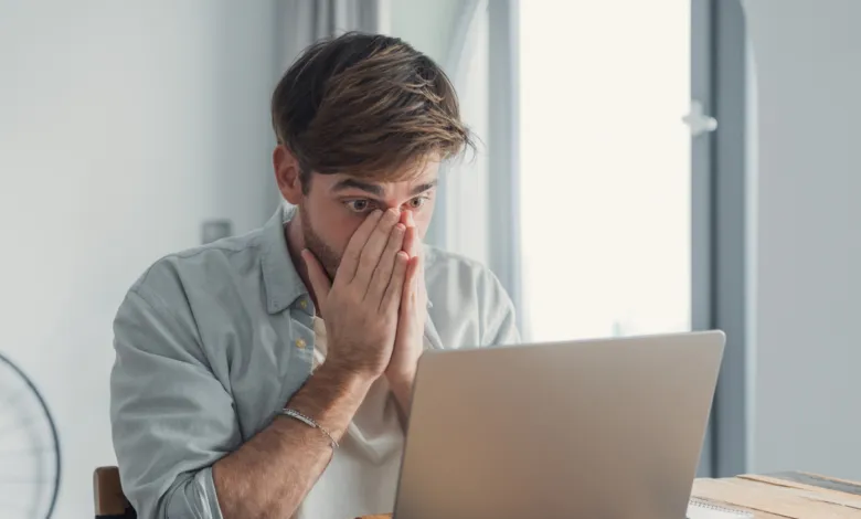 Man with hands on face looking at laptop, appearing stressed or shocked.