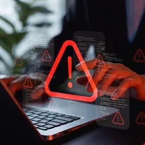 Person types on laptop with red warning sign overlay, indicating a security threat.