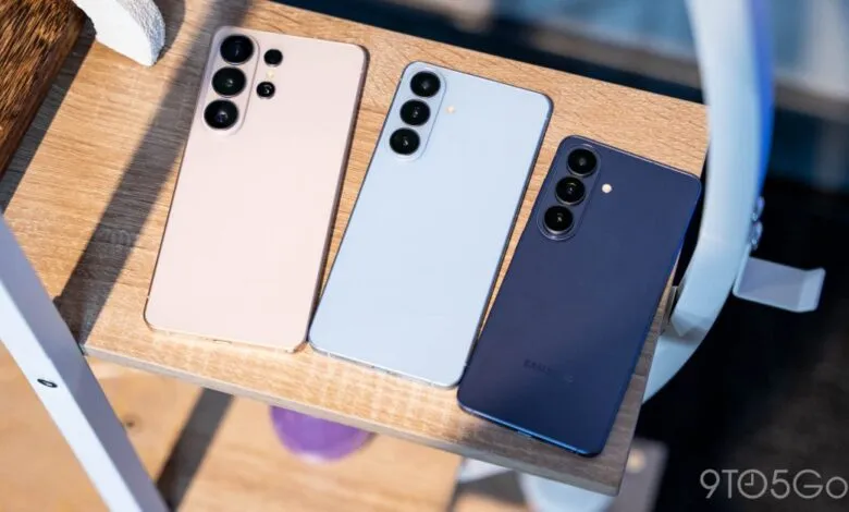 Three Samsung Galaxy S24 phones in pink, light blue, and dark blue on a wooden shelf.