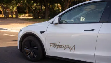 White Tesla car with 'Robotaxi' graphic on the side, driving on a road.