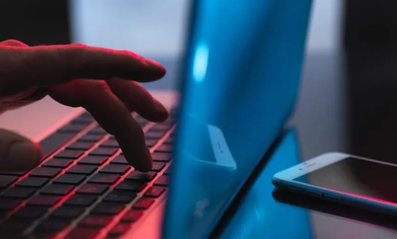 Hand typing on laptop keyboard with smartphone nearby, illuminated in red and blue.