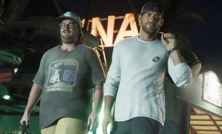 Two men stand in front of a building at night, one wearing a humorous t-shirt.