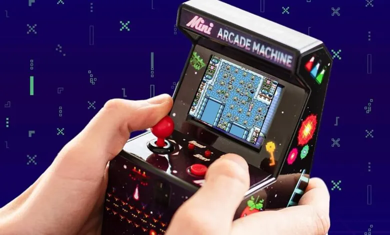 Hands play a mini arcade machine with pixelated game on a blue background.