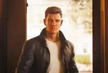 Man in leather jacket stands in doorway, looking forward.