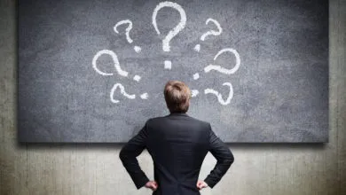 Man in suit faces chalkboard covered in question marks.