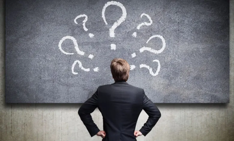 Man in suit faces chalkboard covered in question marks.
