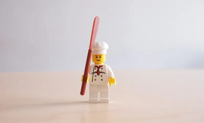 Lego chef minifigure stands holding a spatula against a plain background.