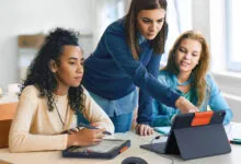 Three young women collaborate on a tablet device at a desk.