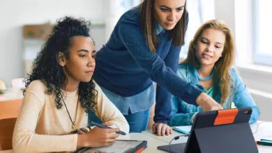 Three young women collaborate on a tablet device at a desk.