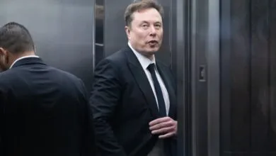 Elon Musk in a suit stands in an elevator, looking forward.