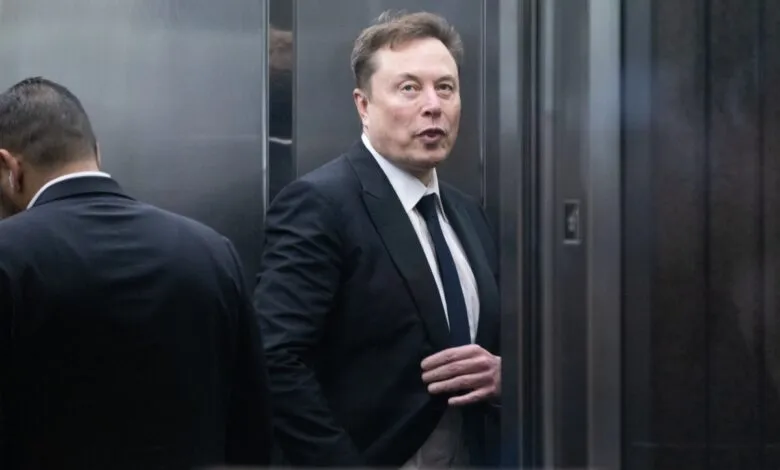 Elon Musk in a suit stands in an elevator, looking forward.