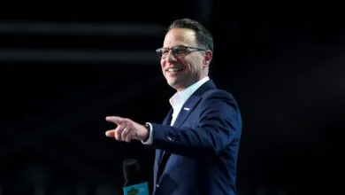 Josh Shapiro smiles, points, and holds a microphone on a dark stage.