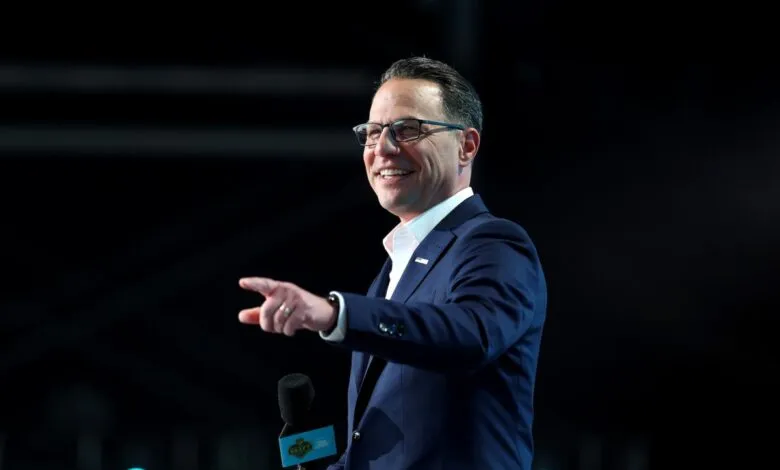 Josh Shapiro smiles, points, and holds a microphone on a dark stage.