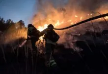 Two firefighters battling a wildfire with a hose at dusk.