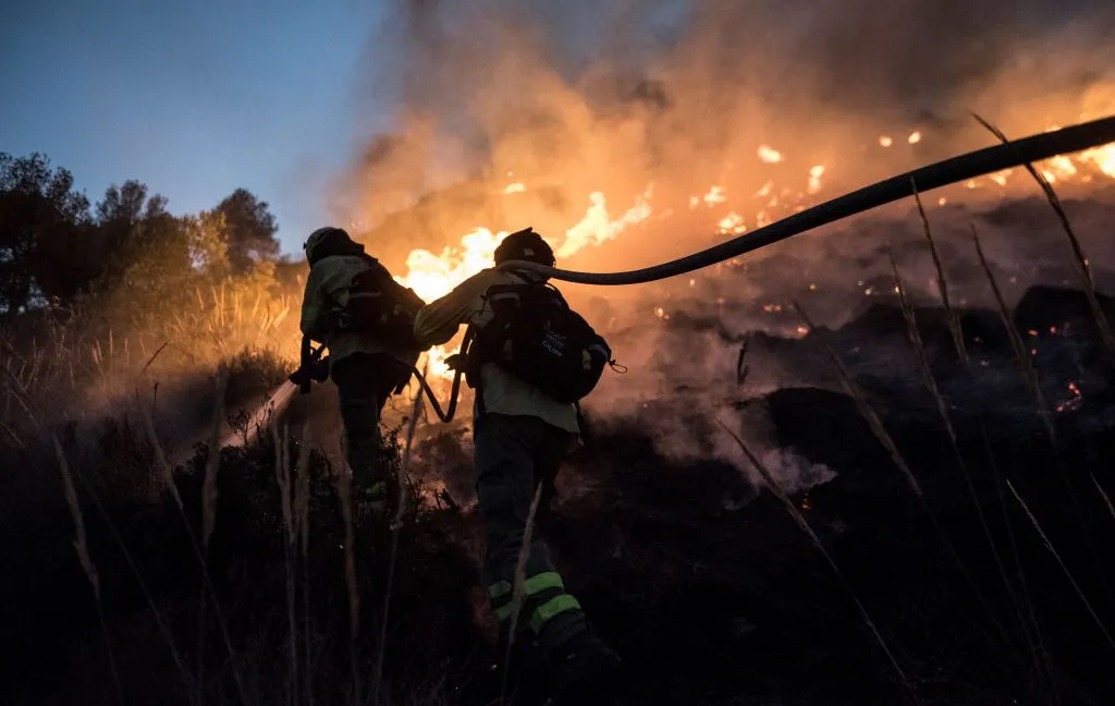 Two firefighters battling a wildfire with a hose at dusk.
