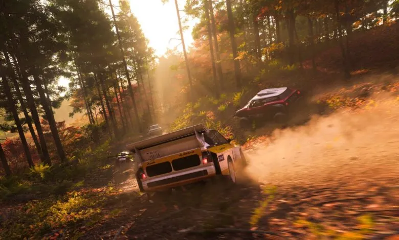 Rally cars race through a sunlit forest on a dirt track, kicking up dust.