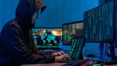 Person in mask and hoodie types on laptop surrounded by monitors with code.