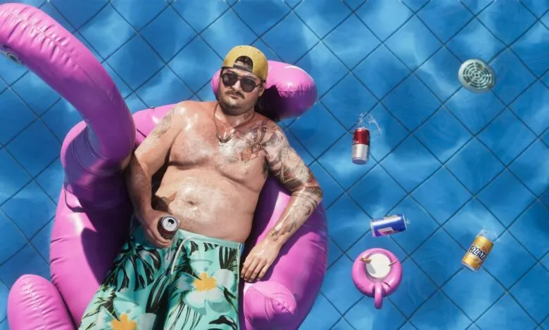 Man relaxing on a pink inflatable flamingo in a bright blue swimming pool with floating beverage cans.
