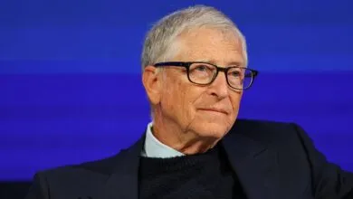 Bill Gates wearing glasses and a dark blazer against a vibrant blue background