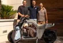 Three men posing with a modern grey electric scooter and a brown dog sitting inside.