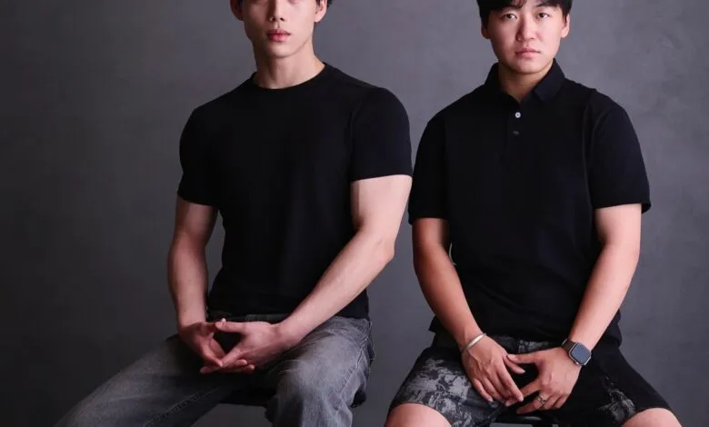 Two young men in black shirts sitting on stools against a neutral gray studio background.