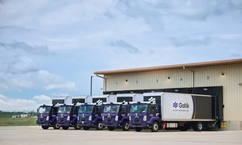 A line of five purple and white Gatik autonomous box trucks parked outside a warehouse.
