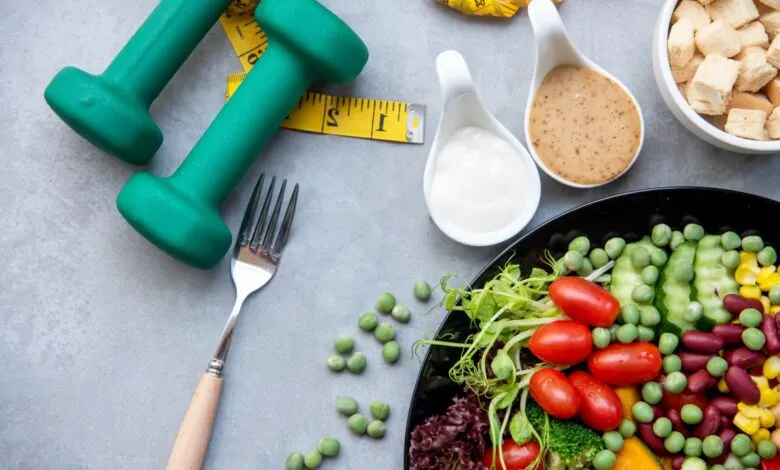 Green dumbbells, a yellow tape measure, and a healthy salad with dressings on a gray surface.