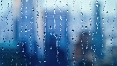 Raindrops and streaks of water run down a glass window against a blurred blue cityscape background
