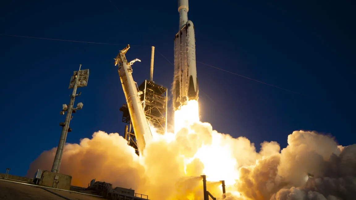 SpaceX Falcon Heavy rocket launching into the night sky with bright orange flames and heavy smoke.