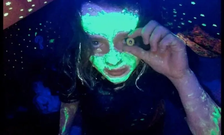 A person with glowing green paint on their face holds up a fake eyeball under blacklight.