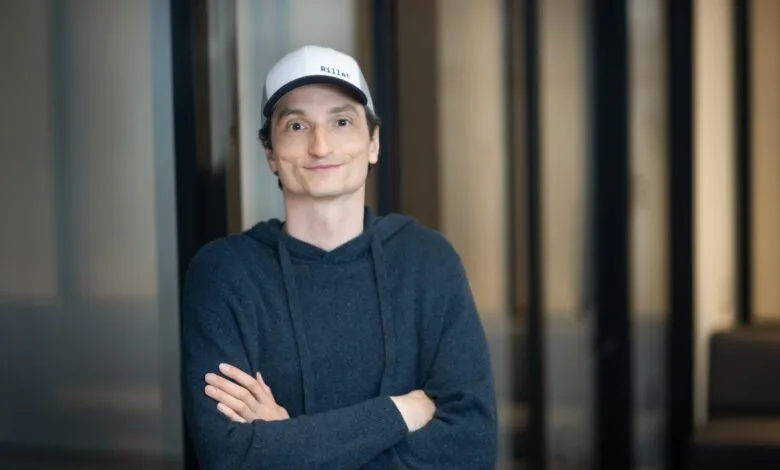 A man wearing a white Rillet cap and dark hoodie with crossed arms smiling in an office setting.