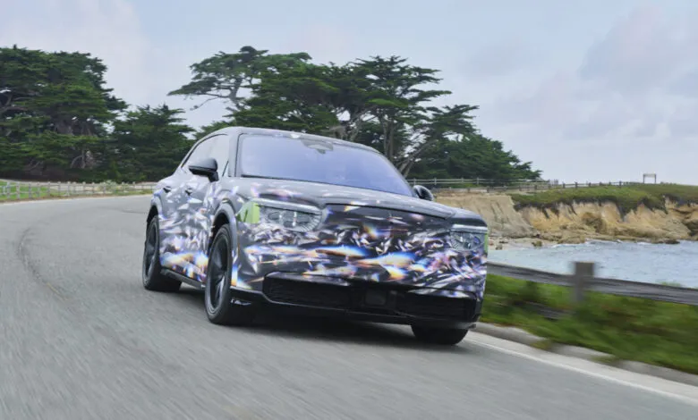 A camouflaged prototype crossover SUV drives along a scenic coastal road with trees and ocean in the background.