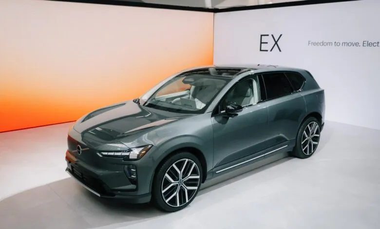 A sleek, dark green Volvo electric SUV on display against a minimalist white and orange backdrop.