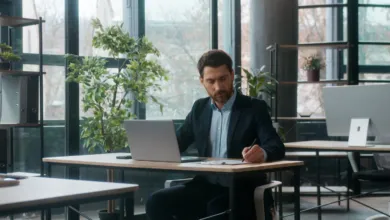 A businessman in a suit sits at a modern office desk, writing notes next to his laptop.