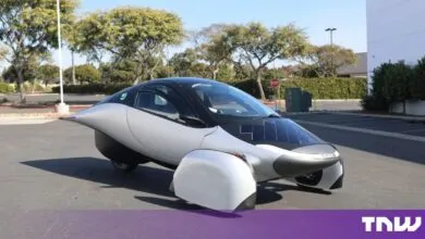 Silver Aptera solar electric vehicle parked outdoors in an asphalt parking lot during the day.