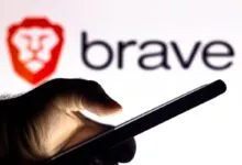 A hand holds a smartphone in front of a screen displaying the Brave browser logo and wordmark.