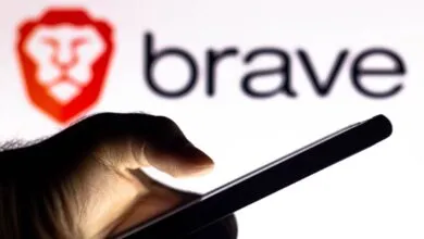 A hand holds a smartphone in front of a screen displaying the Brave browser logo and wordmark.