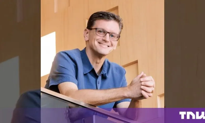 Chris Malone smiling while leaning against a railing in front of a wooden wall.