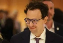Close-up of Alexander Soros wearing dark glasses, a white shirt, and a dark suit jacket indoors.