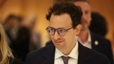Close-up of Alexander Soros wearing dark glasses, a white shirt, and a dark suit jacket indoors.