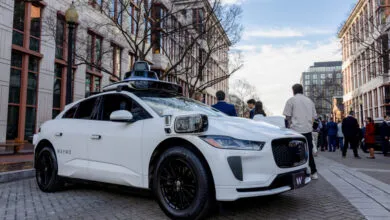 A white Waymo autonomous electric vehicle parked on a city street with pedestrians walking nearby.