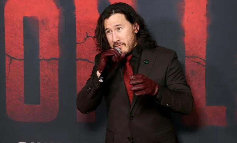Markiplier speaking into a microphone while wearing a brown suit and red leather gloves