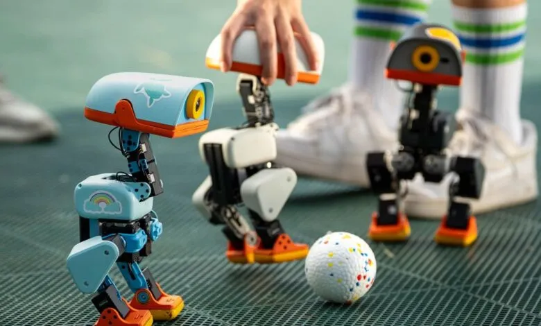 Three colorful small bipedal robots stand near a white ball on a green sports court.