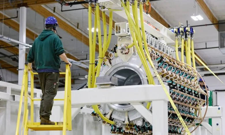 Engineer stands on ladder beside complex cylindrical Pacific Fusion pulser prototype in facility.