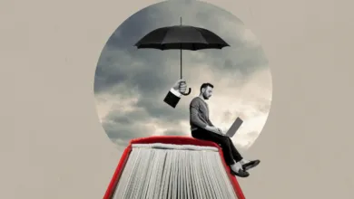A man using a laptop while sitting on a large open red book under a black umbrella.
